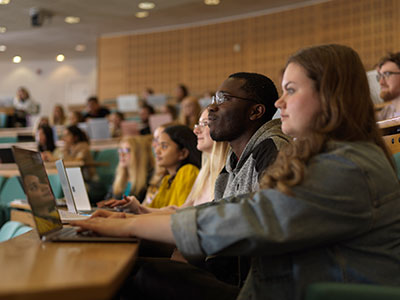 Students sitting in a lecture theatre. The student in the foreground is typing on a laptop.