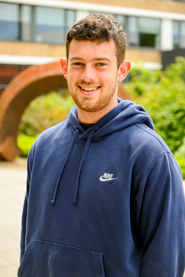 A white man wearing a blue hooded jumper and smiling at the camera on a sunny day.