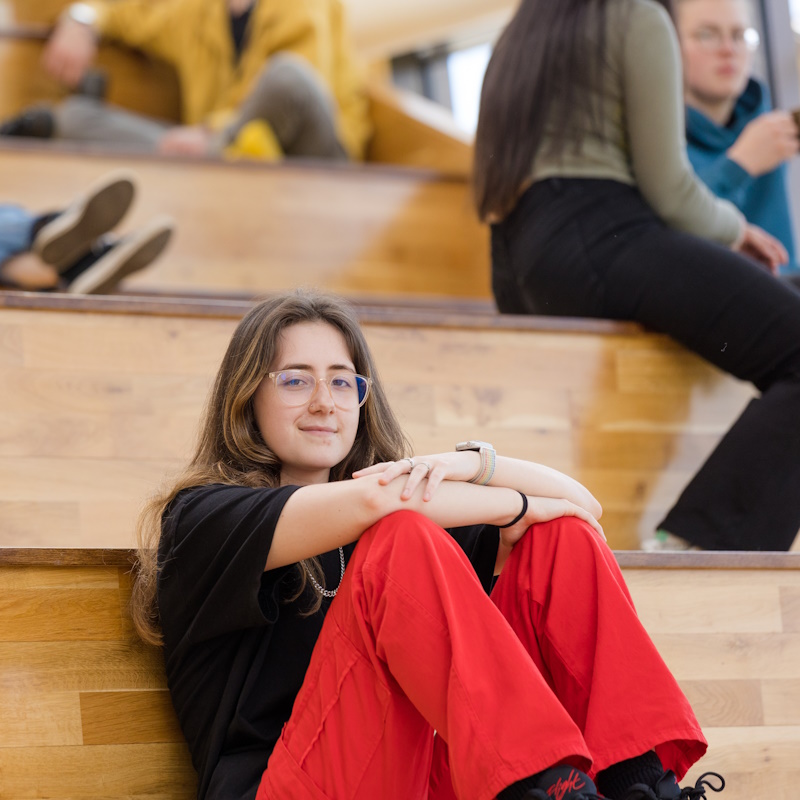 Biology student Katie in black t-shirt and red trousers, sat casually on wooden steps