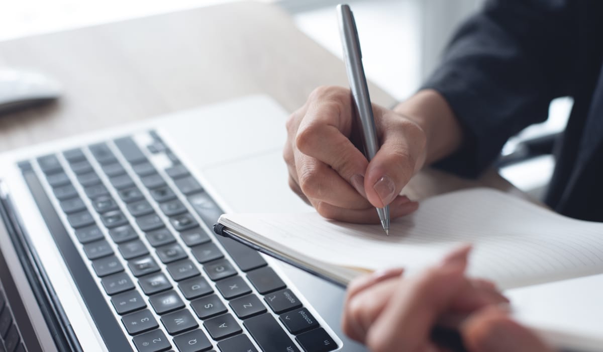 Close up of woman writing on page with pen next to laptop