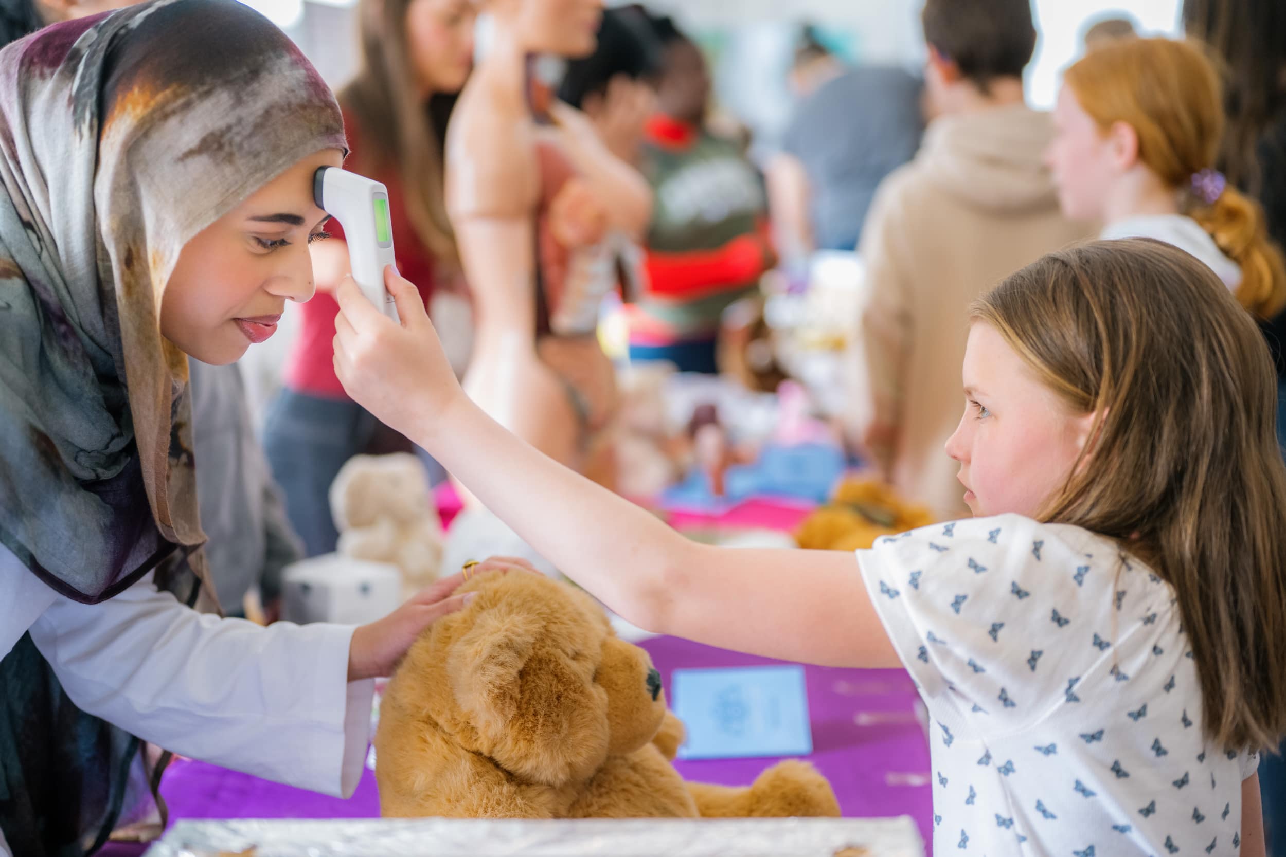 A young child holds a thermometer against the head of a trainee doctor