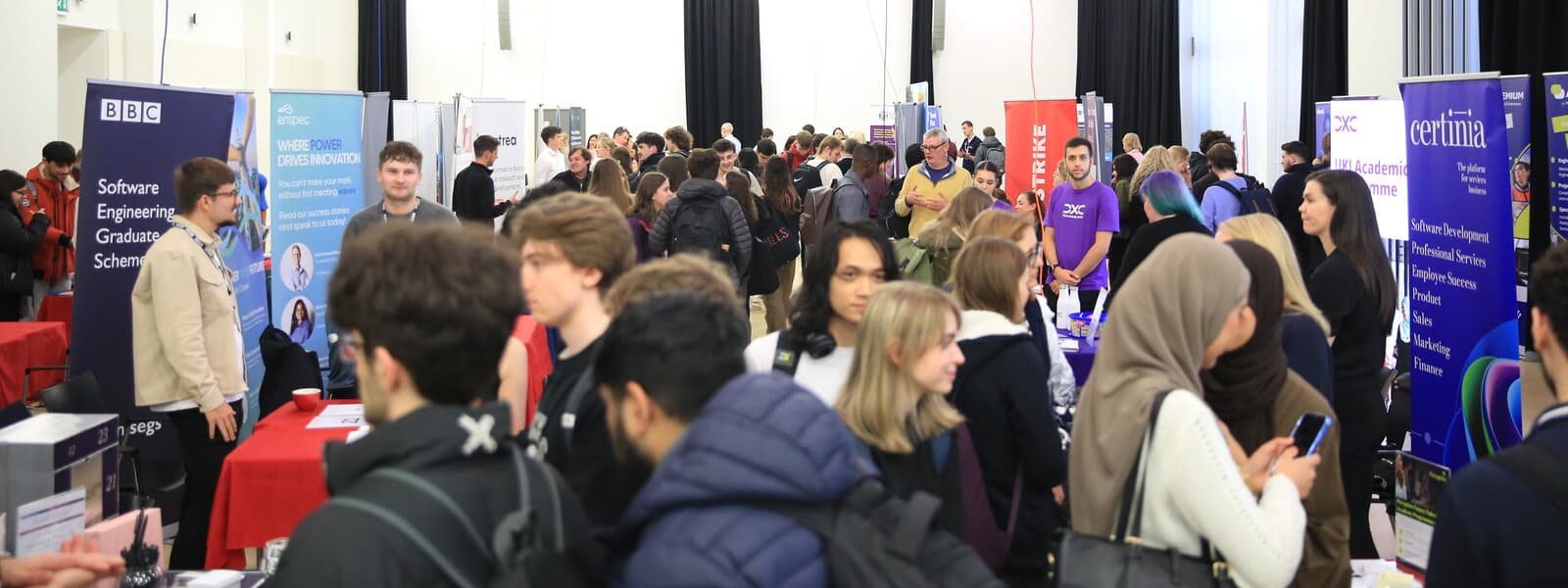 Students speak with an exhibitor at an indoor careers fair, standing beside recruitment display banners in a busy event space.