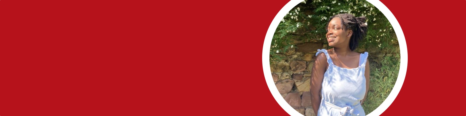 Bethany stands smiling in front of a stone wall with green foliage and flowers dangling down it.