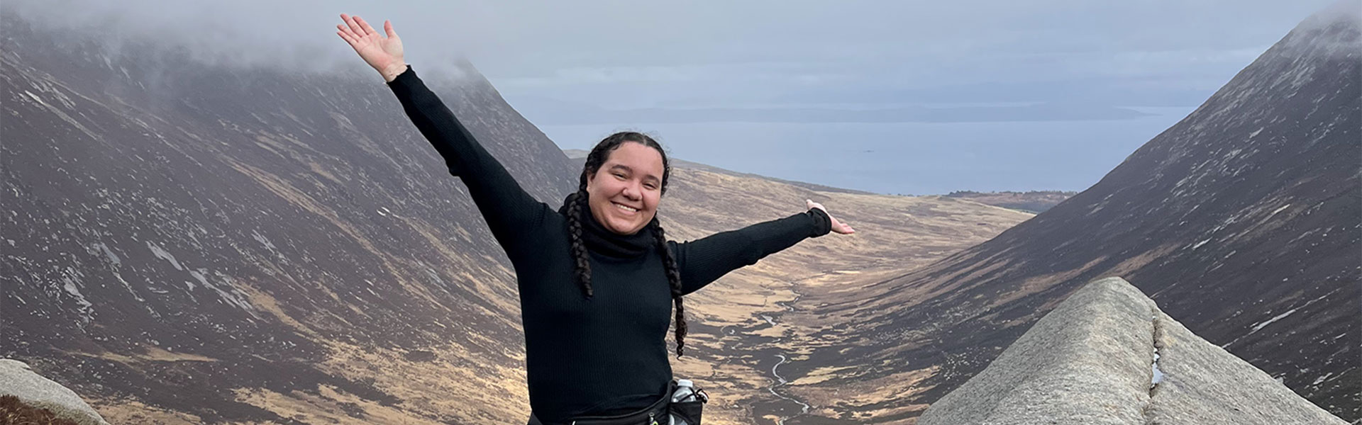 Student Lyea, on a hike surrounded by mountains with arms in the air and big smile on her face