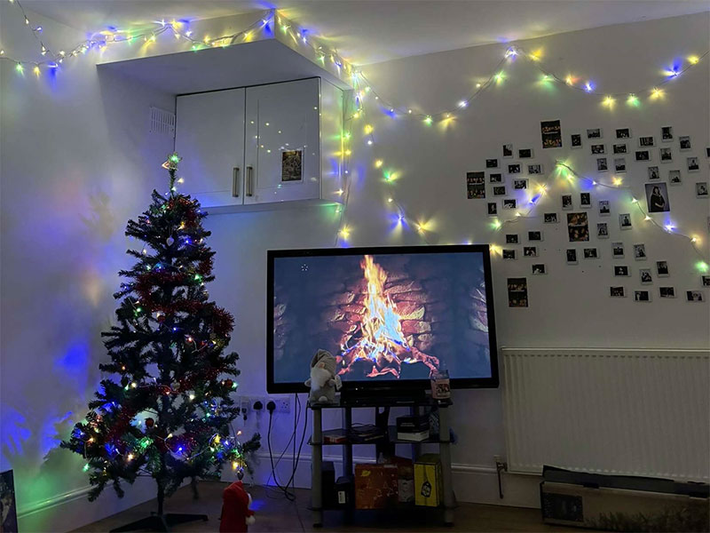 A fireplace displayed on the TV, next to a Christmas tree with Christmas lights in the background.