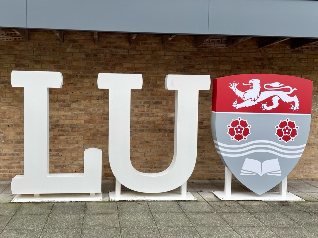 A sign of the letter LU and a red and grey shield of Lancaster university logo.