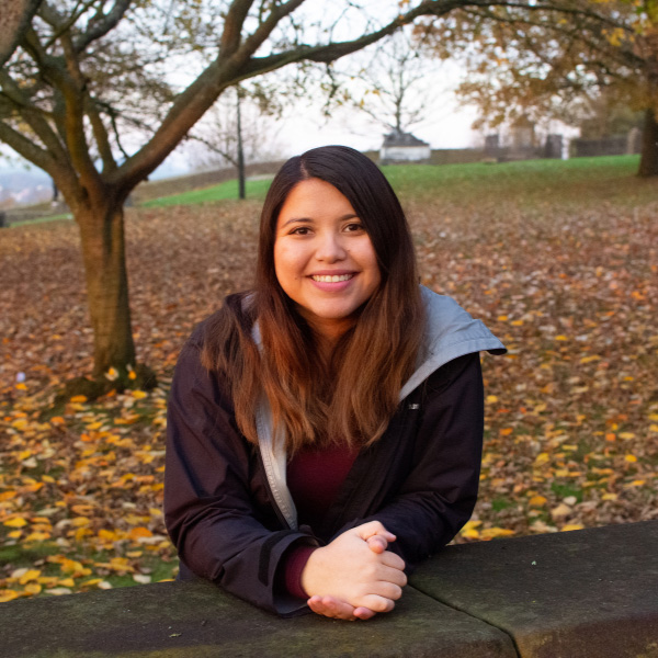 Brenda leaning on a wall and smiling, there are fallen autumn leaves on the grass behind her