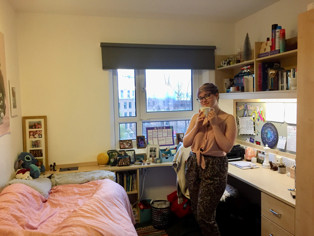 A female student in a bedroom standing drinking a mug of tea