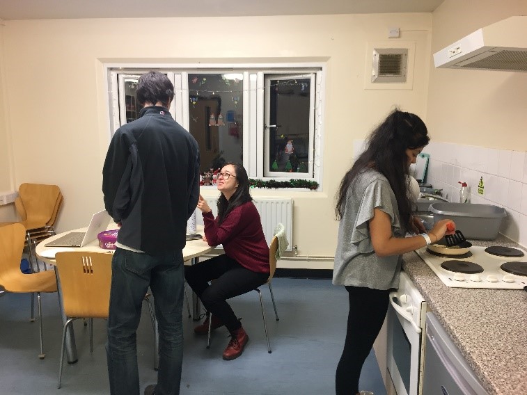 Three students in a campus kitchen, two are chatting at the table and one is cooking at the hob