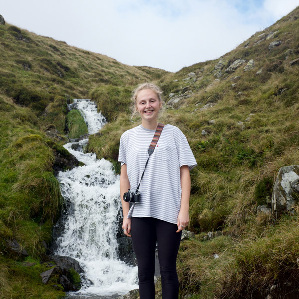 A female student with a camera, standing by a stream looking at the camera