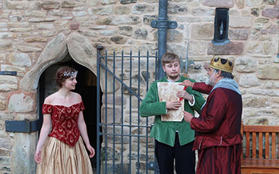 Three students dressed in period clothing performing in front of a stone building.