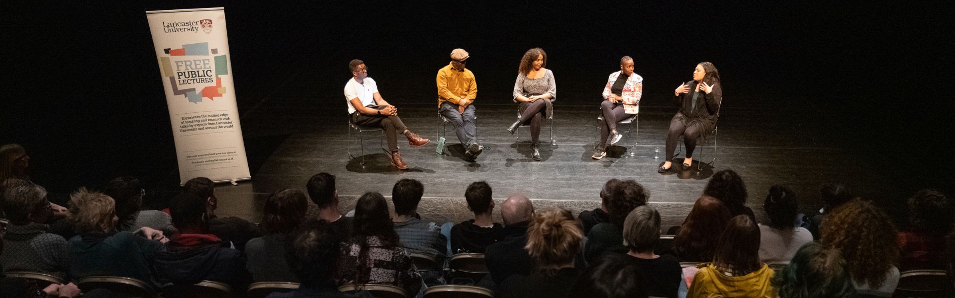a panel of speakers sit on a stage next to a sign saying public lecture