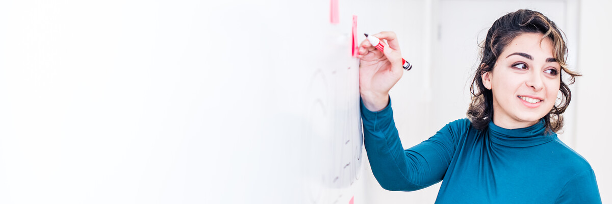 A person writes on a whiteboard, whilst turning around to look at the classroom.