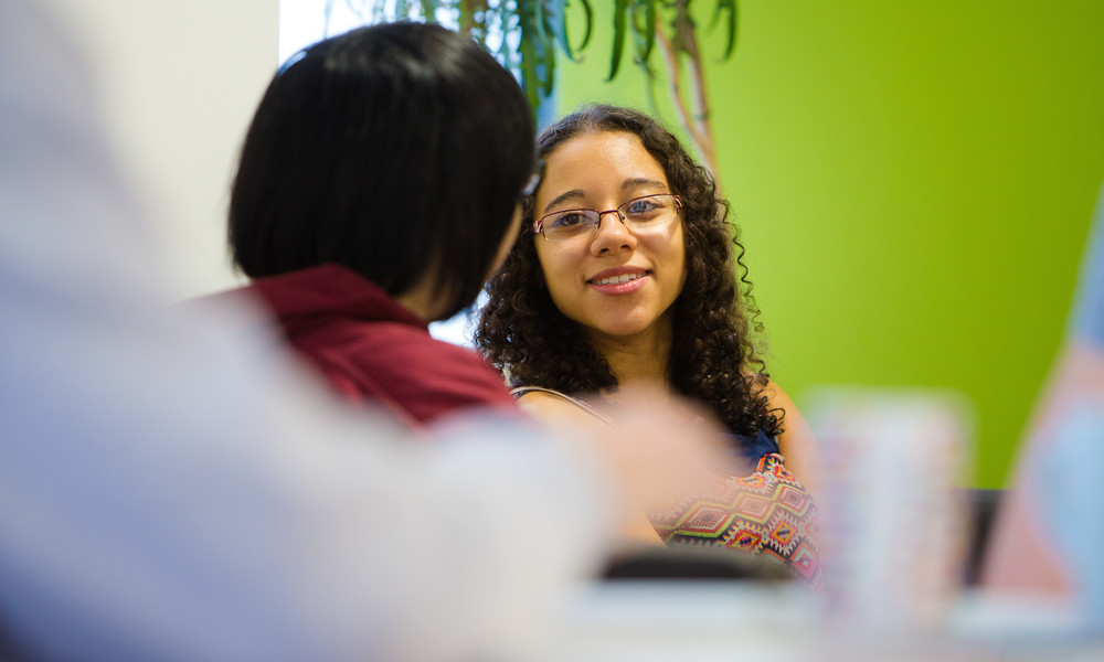 Two students sit chatting in a room with a bright green wall in the background.
