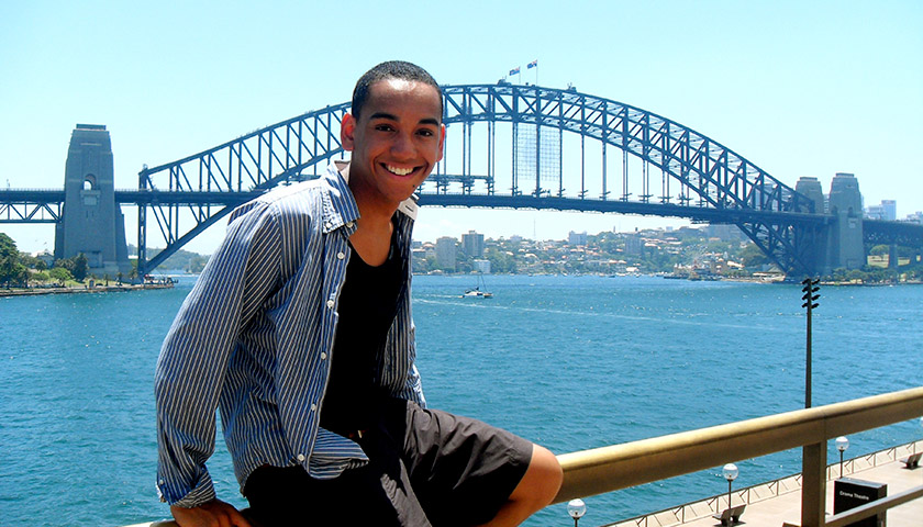 A student sitting in front of a suspension bridge on a sunny day.