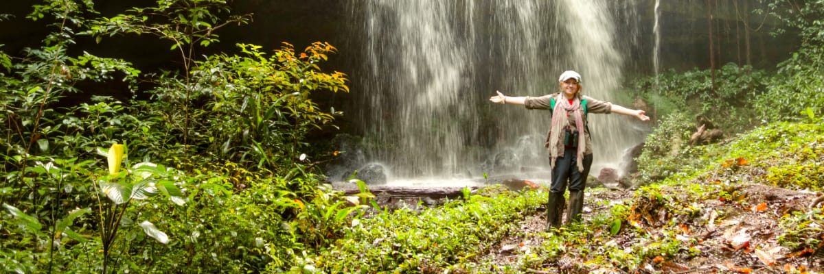 A student stands in front of a waterfall in a heavy jungle in the Amazon.