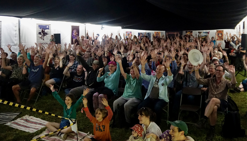 A crowd of people sat in the Physics Pavillion at WOMAD, with their hands raised in the air