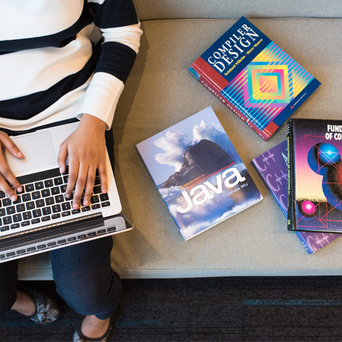 Person sat on a laptop with books on Java and coding next to them