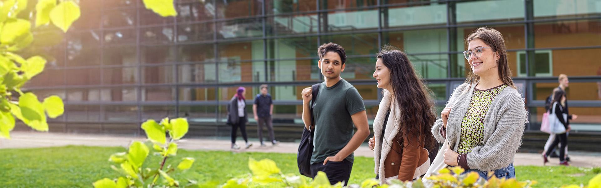 Students walking and chatting in the sunshine with a backdrop of a greenery and modern buildings