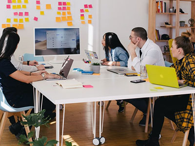 People working on laptops around a white table and looking at a large monitor.