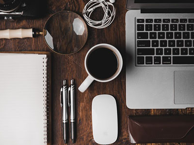 A dark wooden desk with a laptop, keyboard, mouse, notebooks and a cup of coffee.