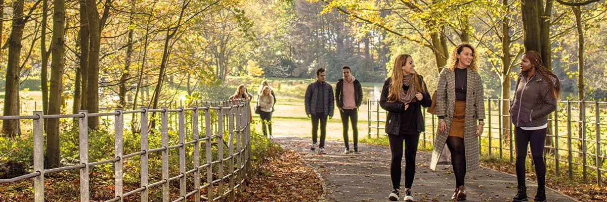 Three people walking along a path in the grounds at Lancaster, with trees and autumn leaves falling.