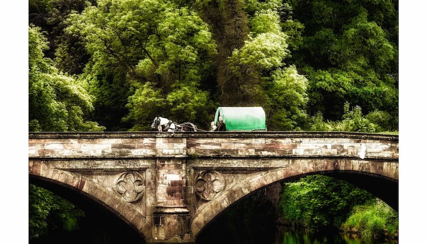 Horse drawn caravan crossing a stone bridge with trees behind.
