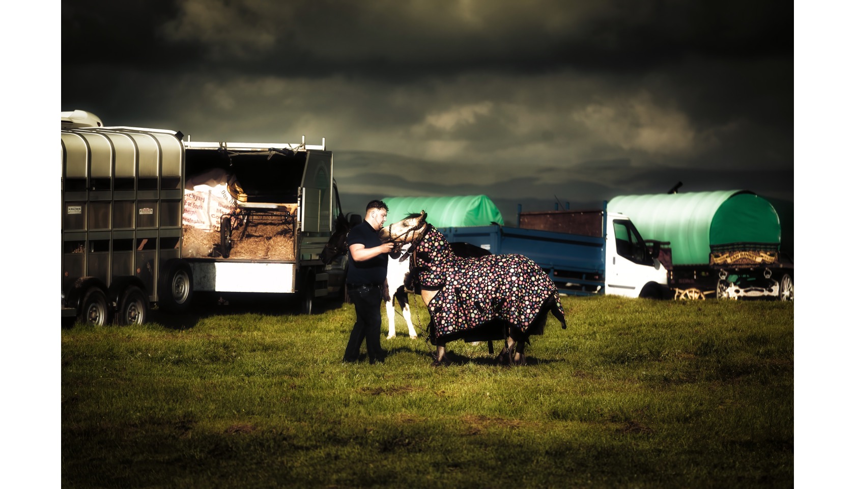 A man holding a horse by the bridle. The horse is wearing a full-length coat. There are caravans.