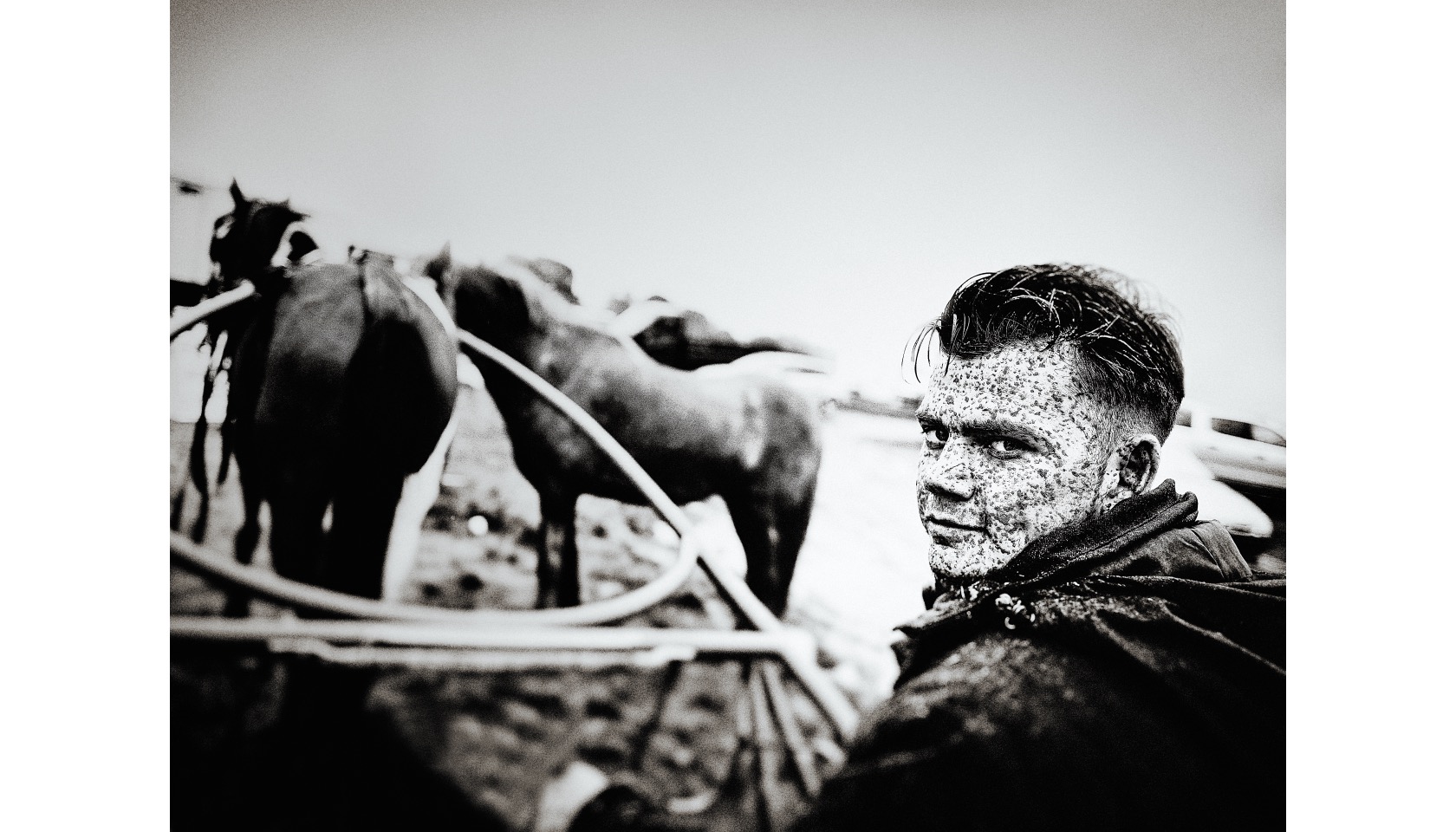 A man with a mud splattered face sitting on a sulky cart with two horses.