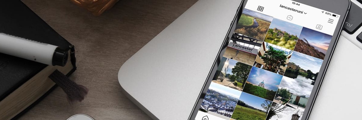 A mobile phone rests on a desk, showing the Lancaster University Instagram feed.