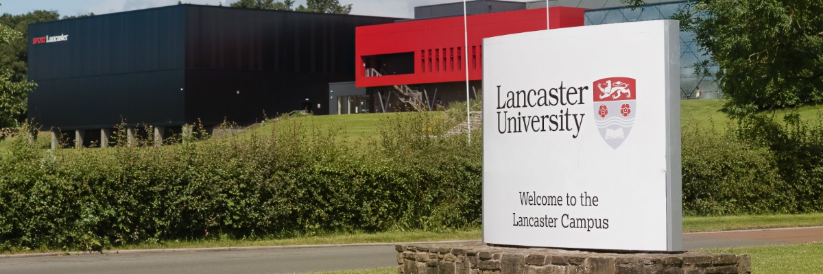 A large white sign welcomes people to Lancaster University. It is surrounded by trees and hedges.