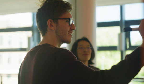 Student writing on a whiteboard with tutor in background