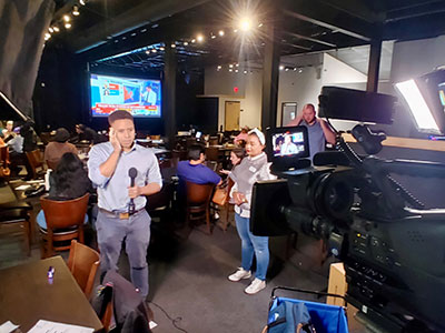 A man surrounded by monitors in front of a professional camera with a microphone.