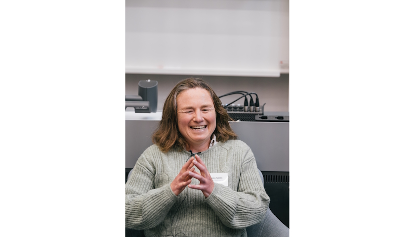 Dr Laura Giles smiling and talking, sitting in front of a grey desk in a lecture theatre.