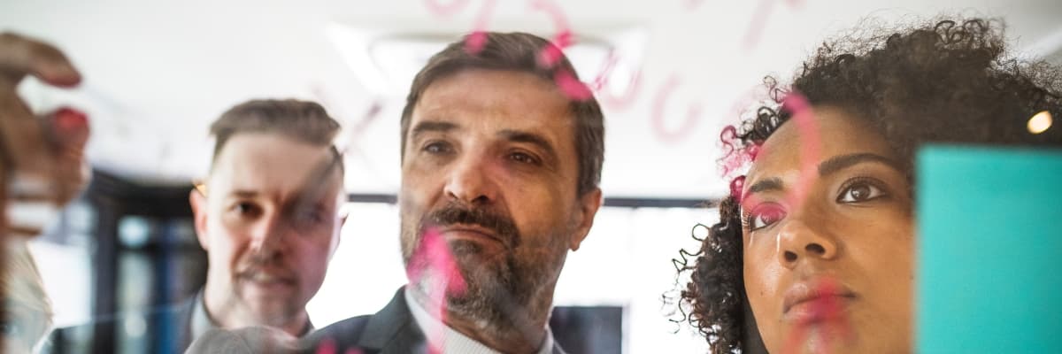 Man writing on perspex wall, flanked by younger man and woman