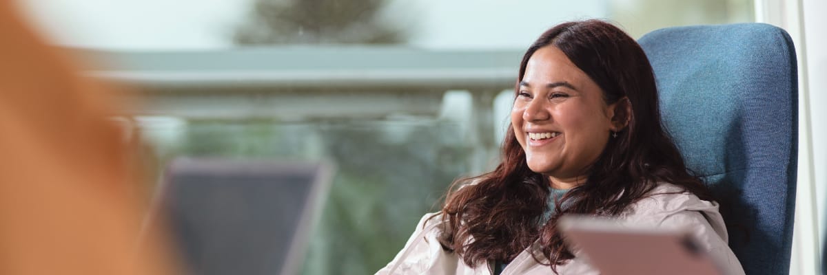A student sits and smiles, with an out of focus landscape outside of the window.