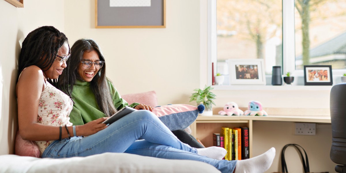 Two students sit, reading side by side on a bed in a brightly lit apartment.