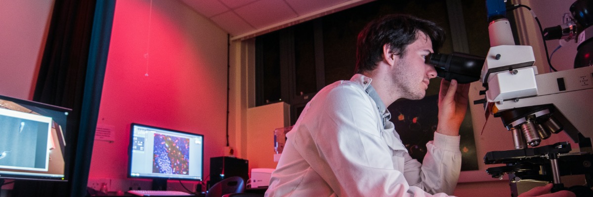A man looks through a microscope in a laboratory lit by red lighting.
