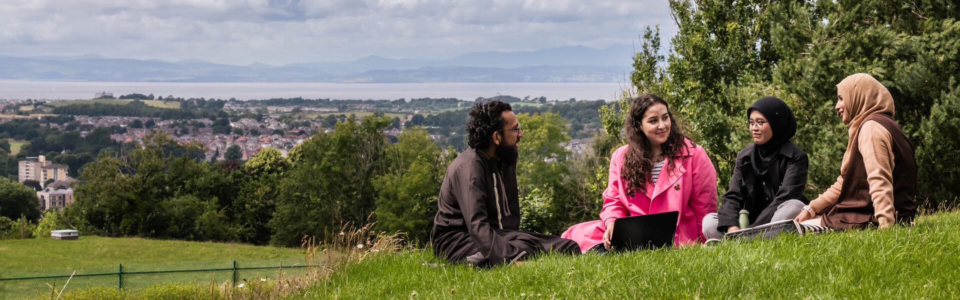 Hero Desktop - Students sitting on a hillside in Williamson Park with the Lake District behind them