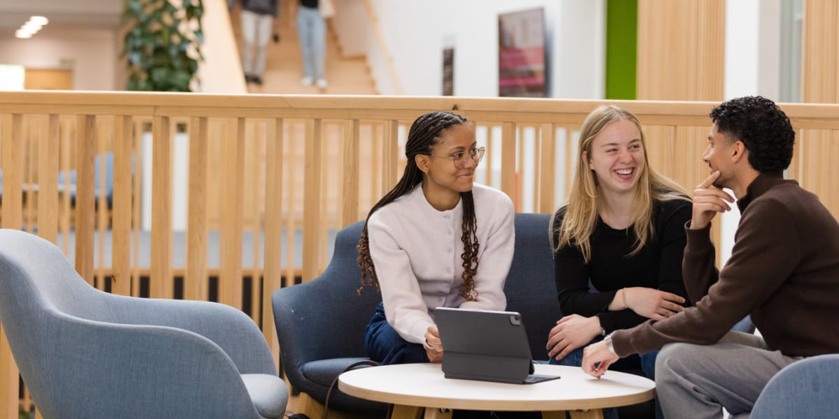 3 students sit around a low table in a bright, open atrium lined with wood.