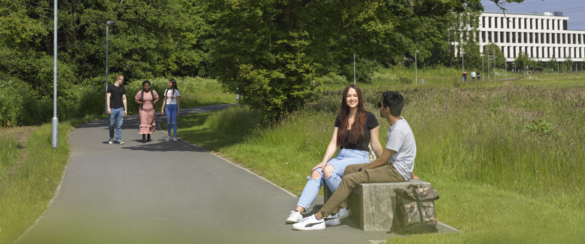 Students on a bench surrounded by greenery. A modernist building is in the background.