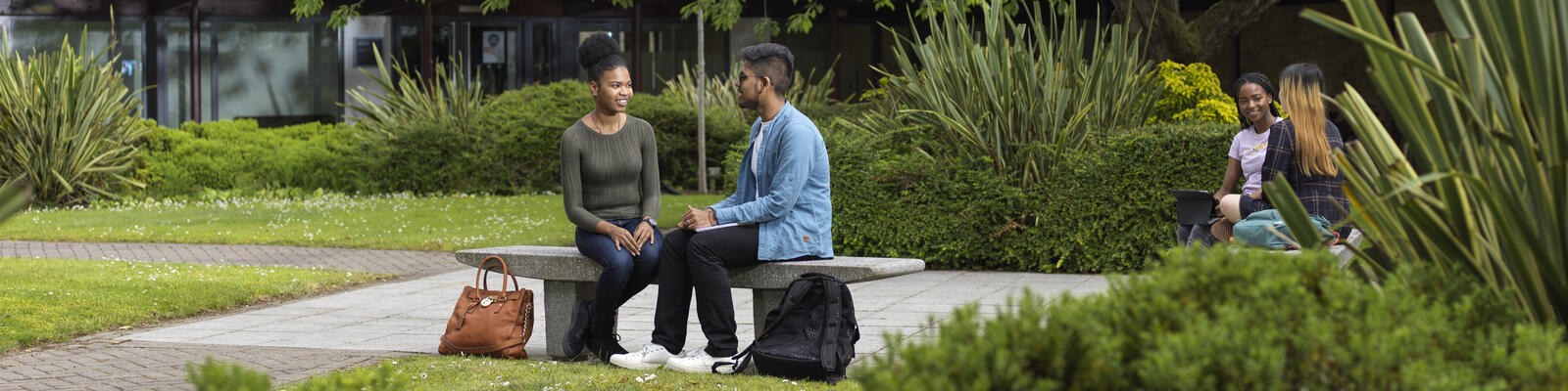 Two students sat on a bench surrounded by trees on campus